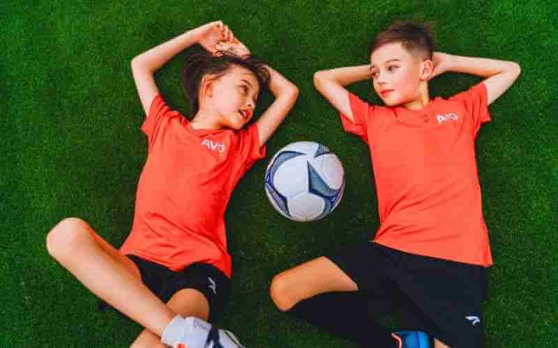 Two kids in 2-color jerseys lying on the artificial turf smiling and staring at each other.